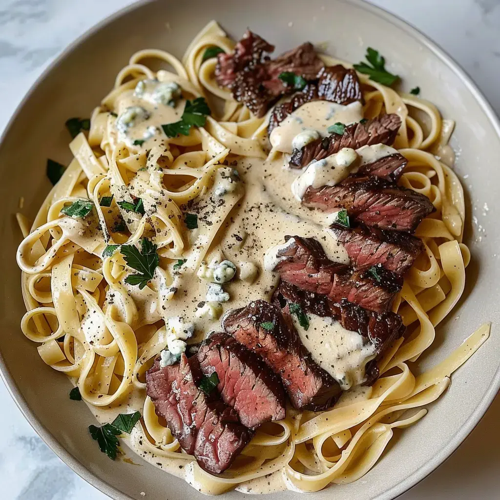 Un plat de pâtes fettuccine servi avec des tranches de steak et une sauce crémeuse, garni de persil et de fromage bleu.