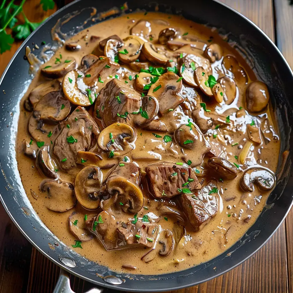 Une poêle contenant des morceaux de viande et des champignons dans une sauce crémeuse, garnie de persil.