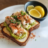 Tranche de pain garnie d'avocat, de poisson et d'épices, servie avec des quartiers de citron.