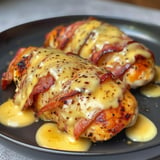 Deux filets de poulet recouverts de fromage fondu et de strips de bacon, servis dans une assiette noire avec une sauce crémeuse.