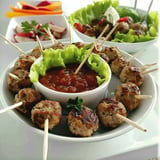 Assiette de boulettes de viande grillées accompagnées d'une sauce dans un bol, le tout servi avec des feuilles de salades et des légumes en garniture.