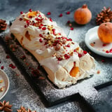 Gâteau roulé à la crème, garni de fruits et de pistaches, décoré de grains de grenade sur une planche noire.