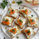 Des tranches de pain en forme d'étoile garnies de fromage frais, de saumon fumé et de persil, présentées sur une assiette décorée de petites étoiles.