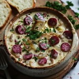 Un bol de soupe crémeuse garni de saucisses, d'oignons et de persil, accompagné de tranches de pain.