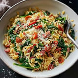 Une assiette de pâtes orzo mélangées avec des épinards, des tomates cerises et du bacon, garnie de fromage râpé.