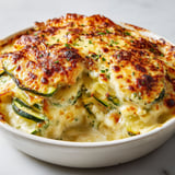 Un plat de gratin savoyard, avec des courgettes et des herbes, est servie sur une table blanche.