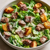 Une salade César revisitée à l'agneau, composée de légumes frais et de viande grillée, est servie dans un bol sur une table.