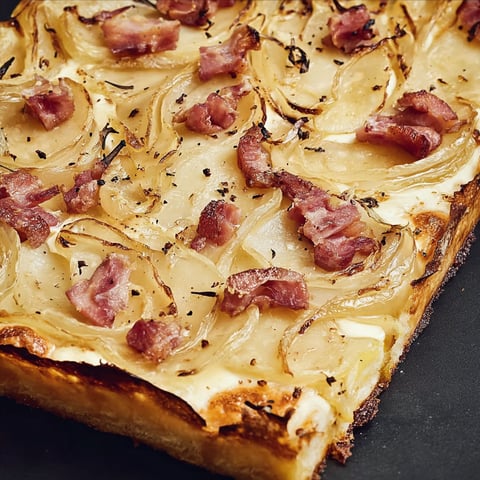 Une flammekueche aux pommes de terre, avec des lardons et des oignons, est servie sur une table noire.